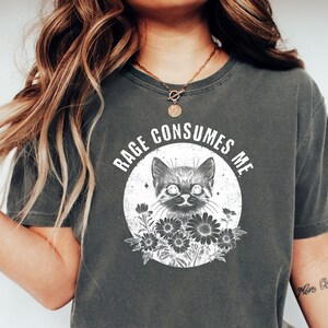 Puede incluir: Camiseta gris oscuro con un gr&aacute;fico desgastado de un gato y flores dentro de un c&iacute;rculo. El texto "RAGE CONSUMES ME" est&aacute; encima del gato. La camiseta es de estilo casual y c&oacute;modo.