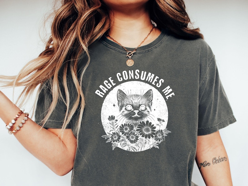 Puede incluir: Camiseta gris oscuro con un gr&aacute;fico desgastado de un gato y flores dentro de un c&iacute;rculo. El texto "RAGE CONSUMES ME" est&aacute; encima del gato. La camiseta es de estilo casual y c&oacute;modo.