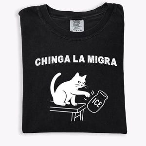 May include: Black t-shirt with white graphic of a cat pushing over an ice jar. The text "CHINGA LA MIGRA" is above the cat illustration. The shirt is folded and has a Comfort Colors tag.