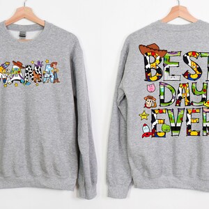 Puede incluir: Sudadera gris con tem&aacute;tica de Toy Story. La parte delantera presenta la palabra "Mama" deletreada con personajes de Toy Story. La parte trasera presenta el texto "Best Day Ever" deletreado con personajes de Toy Story.