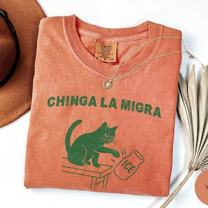 May include: A coral-colored t-shirt with the phrase "CHINGA LA MIGRA" in green block letters. A green cat graphic is depicted knocking over a jar labeled "ICE". A gold necklace and a brown hat are also visible.