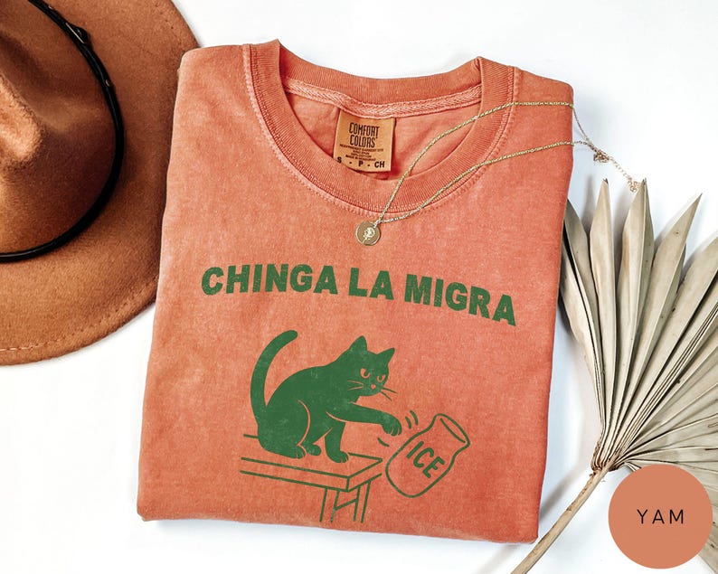 May include: A coral-colored t-shirt with the phrase "CHINGA LA MIGRA" in green block letters. A green cat graphic is depicted knocking over a jar labeled "ICE". A gold necklace and a brown hat are also visible.