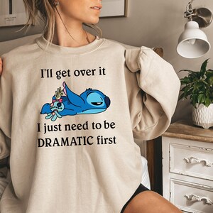 May include: A beige sweatshirt featuring a blue Stitch character from Lilo & Stitch. The text on the sweatshirt reads "I'll get over it I just need to be DRAMATIC first."