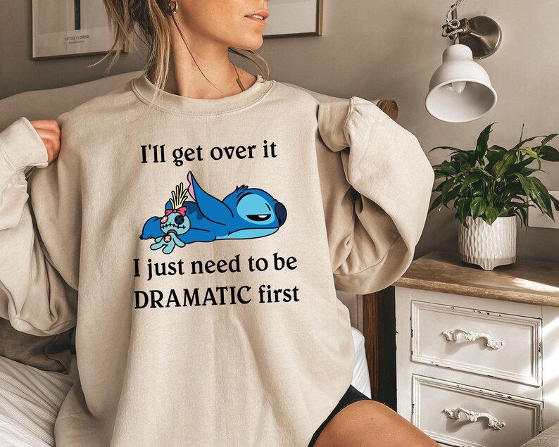 May include: A beige sweatshirt featuring a blue Stitch character from Lilo & Stitch. The text on the sweatshirt reads "I'll get over it I just need to be DRAMATIC first."
