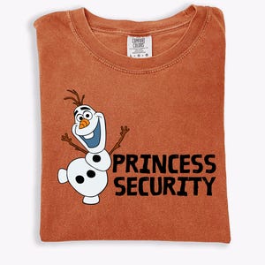 May include: A rust-colored t-shirt featuring a cartoon image of Olaf the snowman with the text "PRINCESS SECURITY" in black. The shirt is folded and has a Comfort Colors tag.