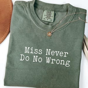 May include: An olive green t-shirt with the text "Miss Never Do No Wrong" in white. A gold necklace with a circular pendant rests on the shirt. A brown hat and dried plant are in the background.