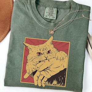 May include: Sage green t-shirt featuring a graphic of an angry cat biting a hand, set within a red and gold frame. A silver necklace with a small pendant rests on the shirt. The shirt is a Comfort Colors brand, size S.