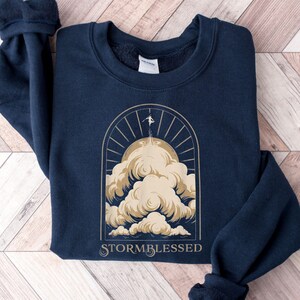 May include: Navy blue sweatshirt with a gold graphic of a person standing on a cloud with the text "STORMBLESSED" underneath.