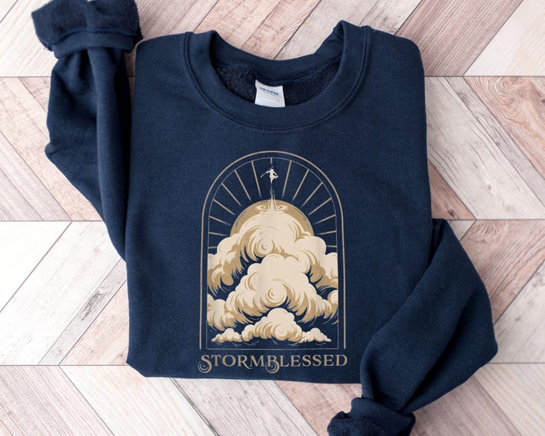 May include: Navy blue sweatshirt with a gold graphic of a person standing on a cloud with the text "STORMBLESSED" underneath.