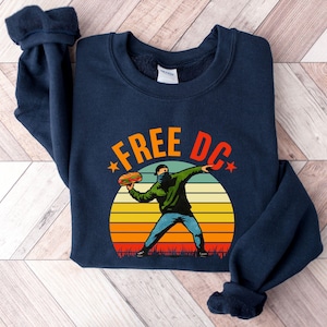 May include: Navy blue sweatshirt with the text "FREE DC" in orange and a graphic of a person throwing a hot dog. The design includes a sunset background with orange, yellow, and green stripes.