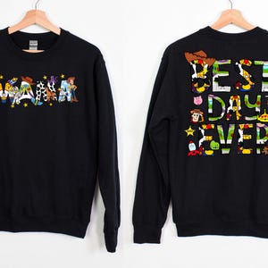 Puede incluir: Sudadera negra con tem&aacute;tica de Toy Story. La parte delantera presenta la palabra "WAMA" deletreada con personajes de Toy Story. La parte trasera presenta el texto "BEST DAY EVER" deletreado con personajes de Toy Story.