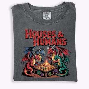 May include: Dark gray t-shirt with the text "HOUSES & HUMANS" in red. The graphic features four dragons of different colors gathered around a table with a bonfire. The shirt is folded on a white surface.