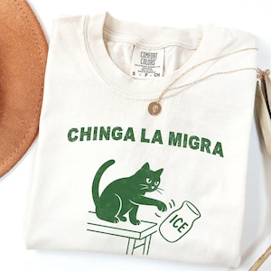 May include: Cream-colored t-shirt with the text "CHINGA LA MIGRA" in green. A green cat illustration is depicted knocking over an ice jug. A gold necklace and a brown hat are also visible.