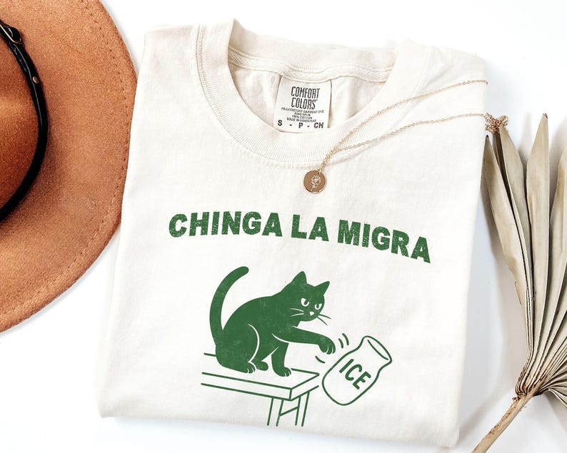 May include: Cream-colored t-shirt with the text "CHINGA LA MIGRA" in green. A green cat illustration is depicted knocking over an ice jug. A gold necklace and a brown hat are also visible.