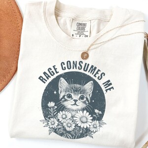 Puede incluir: Camiseta color crema con la frase "RAGE CONSUMES ME" que rodea un gr&aacute;fico de un gatito y margaritas. Un collar dorado con un peque&ntilde;o colgante est&aacute; sobre la camiseta.