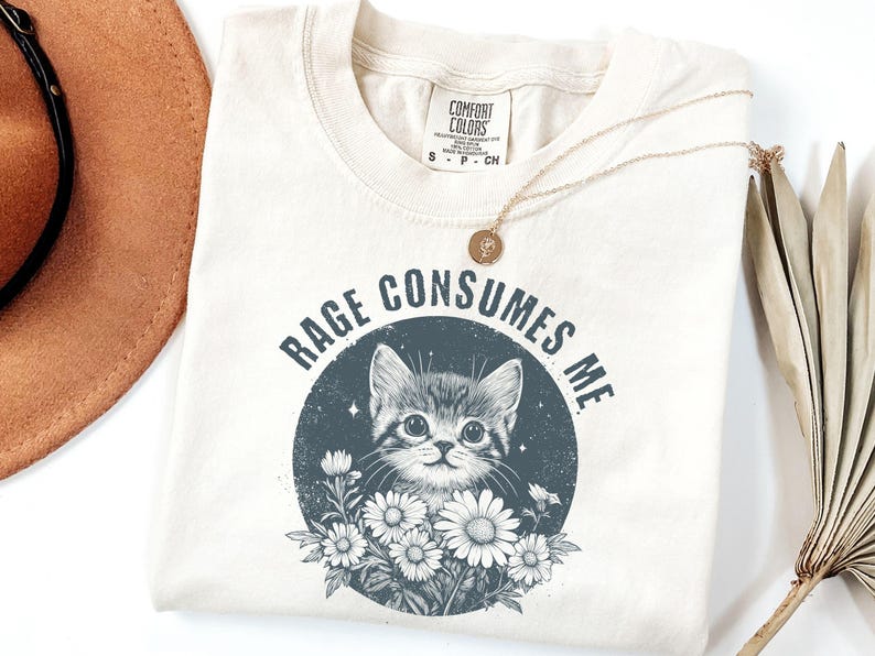 Puede incluir: Camiseta color crema con la frase "RAGE CONSUMES ME" que rodea un gr&aacute;fico de un gatito y margaritas. Un collar dorado con un peque&ntilde;o colgante est&aacute; sobre la camiseta.