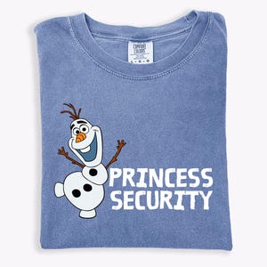May include: A blue t-shirt with a graphic of Olaf the snowman and the words "PRINCESS SECURITY" in white. The shirt is folded and has a Comfort Colors tag at the neckline.