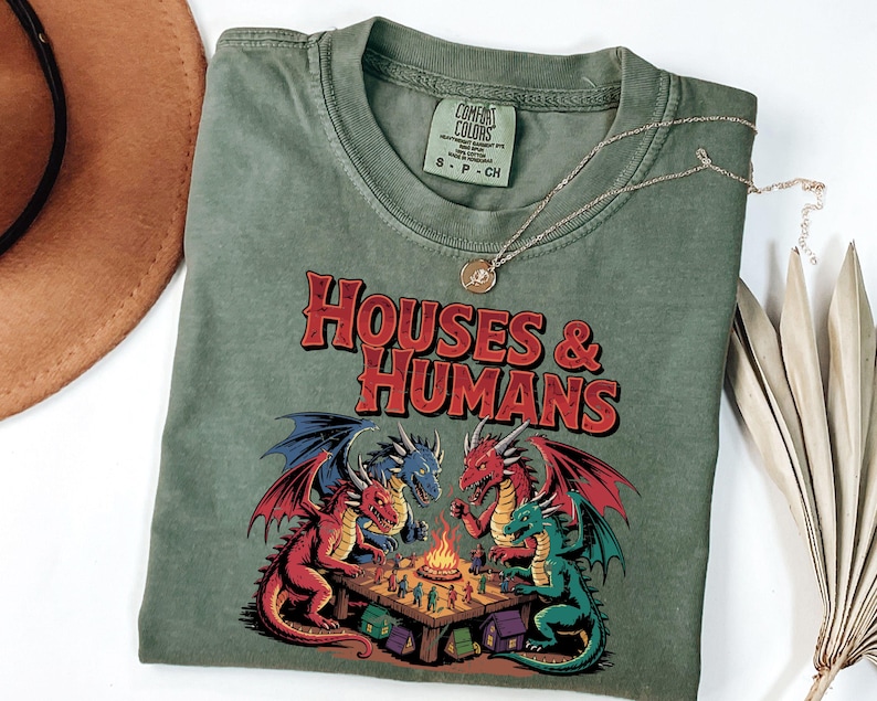 May include: A sage green t-shirt with the text "HOUSES & HUMANS" in red. The graphic features dragons around a campfire. A gold necklace is draped across the shirt. A brown hat and dried plant are in the background.