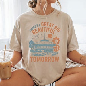 May include: Beige t-shirt with the text "There's a Great Big Beautiful Tomorrow" in orange and blue. The design includes a futuristic building, sun, clouds, and gears. The shirt is a relaxed fit.
