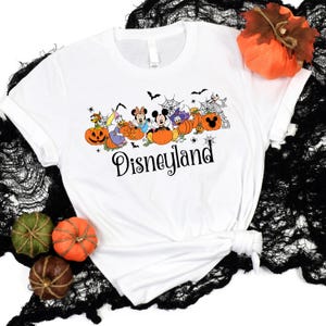May include: White t-shirt with a Halloween-themed design featuring Mickey Mouse and other Disney characters dressed in costumes and holding pumpkins. The text "Disneyland" is printed below the characters.
