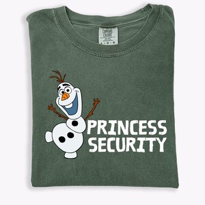May include: A green Comfort Colors t-shirt featuring a cartoon image of Olaf the snowman with the words "PRINCESS SECURITY" in white block letters. The shirt is folded and has a tag at the neckline.