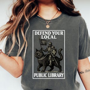 May include: Dark gray t-shirt with a graphic featuring a knight riding a black cat, holding a flag. The text "DEFEND YOUR LOCAL PUBLIC LIBRARY" is printed in a white rectangular frame. The design is vintage-inspired.