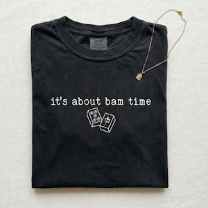 May include: Black t-shirt with the phrase "it's about bam time" and two white graphic illustrations of mahjong tiles. A gold necklace with a small pendant rests on the shirt.
