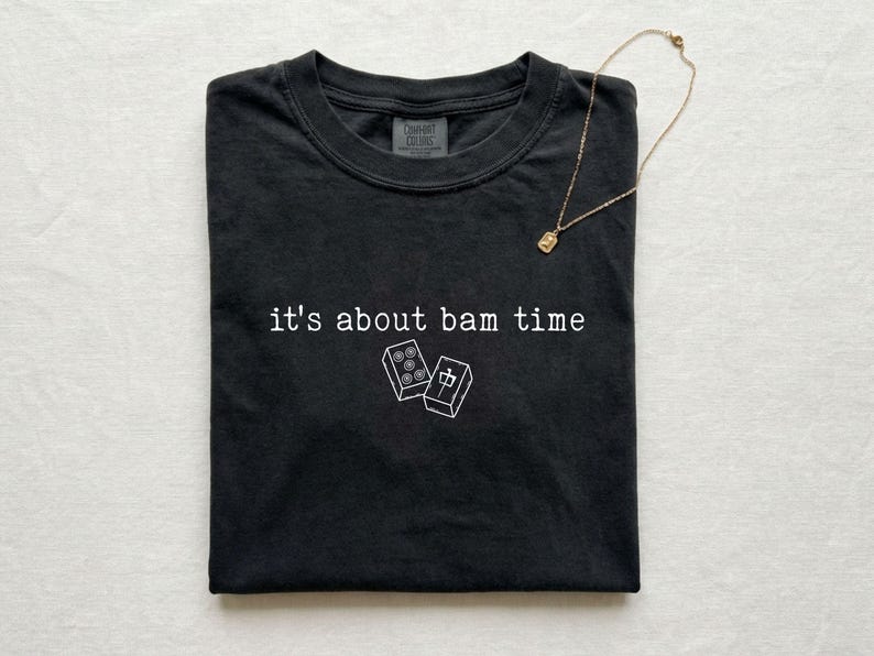 Pu&ograve; includere: T-shirt nero con la scritta "it's about bam time" e due illustrazioni grafiche bianche di tessere di mahjong. Una collana d'oro con un piccolo ciondolo &egrave; appoggiata sulla maglietta.