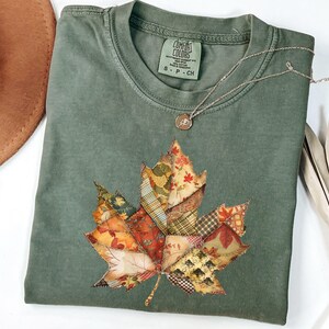 May include: Sage green t-shirt featuring a patchwork leaf design in autumn colors. The leaf is made of various fabric patterns. A delicate silver necklace with a small pendant rests on the shirt. The tag reads "Comfort Colors".