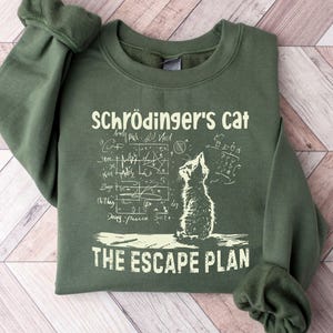 May include: A green sweatshirt with the text "Schrödinger's Cat" above a cat illustration and mathematical equations. Below the cat is the text "The Escape Plan". The sweatshirt has long sleeves and a crew neck.