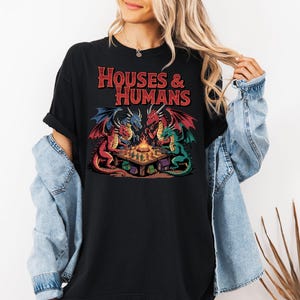 May include: Black t-shirt featuring a graphic of three dragons around a table with a campfire. The dragons are red, blue, and green with detailed wings. The text "HOUSES & HUMANS" is in red, stylized lettering.