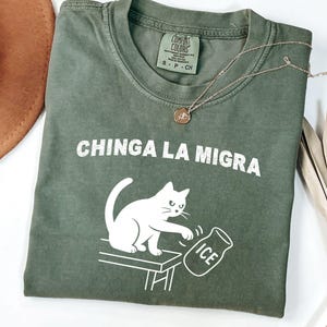 May include: An olive green t-shirt with the phrase "CHINGA LA MIGRA" in white block letters. A white cat illustration is featured, knocking over an ice jug. A gold necklace and a brown hat are also visible.
