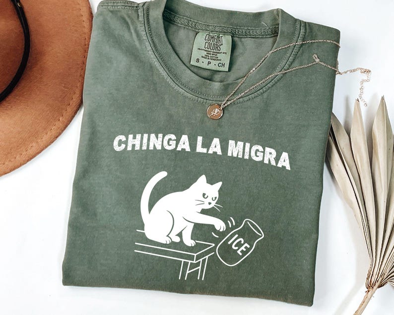 May include: An olive green t-shirt with the phrase "CHINGA LA MIGRA" in white block letters. A white cat illustration is featured, knocking over an ice jug. A gold necklace and a brown hat are also visible.
