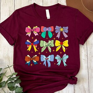 May include: A maroon t-shirt with a graphic of nine colorful bows. The bows are arranged in a grid pattern and have different designs and colors, including pink, yellow, green, blue, and red.