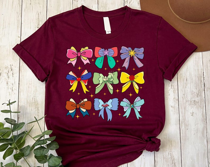 May include: A maroon t-shirt with a graphic of nine colorful bows. The bows are arranged in a grid pattern and have different designs and colors, including pink, yellow, green, blue, and red.