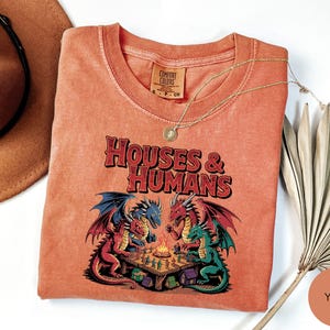 May include: A coral-colored t-shirt with the text "HOUSES & HUMANS" above a graphic of dragons playing a board game. The shirt has a crew neck and a small tag on the collar. A gold necklace and a hat are next to the shirt.