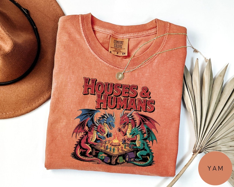 May include: A coral-colored t-shirt with the text "HOUSES & HUMANS" above a graphic of dragons playing a board game. The shirt has a crew neck and a small tag on the collar. A gold necklace and a hat are next to the shirt.