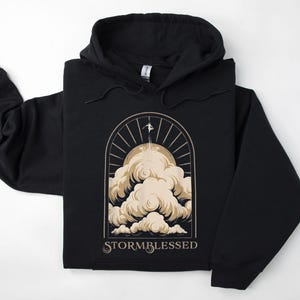 May include: Black hooded sweatshirt with a graphic of a person standing on a cloud with the text "STORMBLESSED" below.