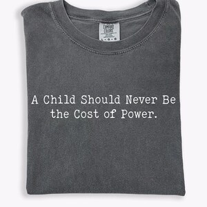 Puede incluir: Camiseta gris oscuro con el texto blanco "A Child Should Never Be the Cost of Power". La camiseta est&aacute; doblada y tiene cuello redondo. La etiqueta de la marca Comfort Colors es visible.