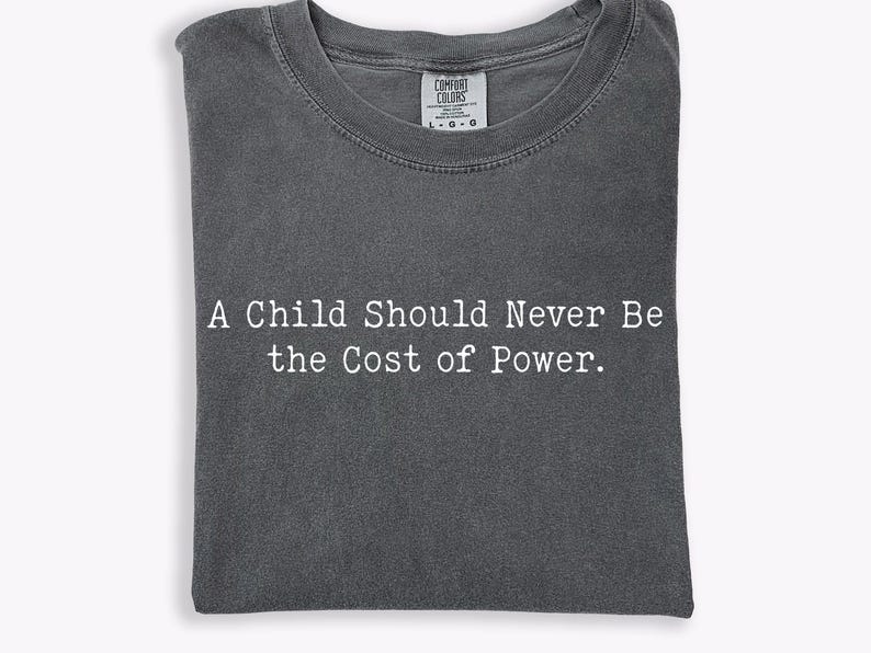 Puede incluir: Camiseta gris oscuro con el texto blanco "A Child Should Never Be the Cost of Power". La camiseta est&aacute; doblada y tiene cuello redondo. La etiqueta de la marca Comfort Colors es visible.