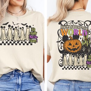 May include: Beige t-shirt with a Halloween-themed graphic. The front features cartoon ghosts and the text "It's really Not-So-Scary!". The back displays a jack-o'-lantern, castle, and the words "Haunt Around The World".