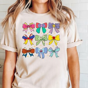 May include: A beige t-shirt with a colorful bow design. The bows are in various colors, including pink, blue, green, yellow, and red. The bows are arranged in a grid pattern on the front of the shirt.