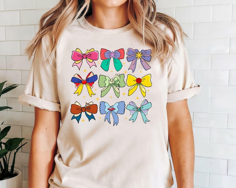 May include: A beige t-shirt with a colorful bow design. The bows are in various colors, including pink, blue, green, yellow, and red. The bows are arranged in a grid pattern on the front of the shirt.