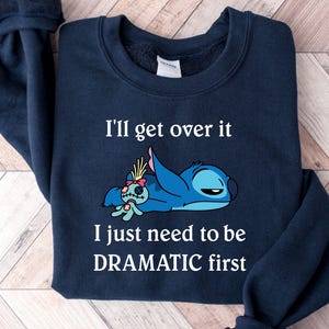 May include: A navy blue sweatshirt featuring a white graphic of Stitch from Lilo & Stitch. The graphic depicts Stitch lying down with a sad expression and the text "I'll get over it I just need to be DRAMATIC first".