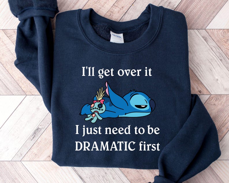 May include: A navy blue sweatshirt featuring a white graphic of Stitch from Lilo & Stitch. The graphic depicts Stitch lying down with a sad expression and the text "I'll get over it I just need to be DRAMATIC first".