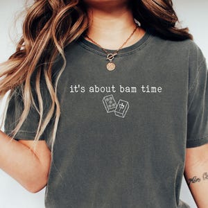 Pu&ograve; includere: T-shirt grigio scuro con la scritta bianca "it's about bam time" e una piccola grafica di domino. Una t-shirt casual e unisex.