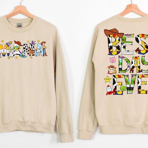 Puede incluir: Una sudadera de color beige con tem&aacute;tica de Toy Story. La parte delantera de la sudadera dice "Mama" con personajes de Toy Story. La parte trasera de la sudadera dice "Best Day Ever" con personajes de Toy Story.