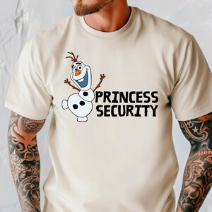 May include: Beige t-shirt featuring a cartoon image of Olaf from Frozen, with the text "PRINCESS SECURITY" in black. Olaf is white with a carrot nose, twig arms, and a brown hair. The shirt has short sleeves.