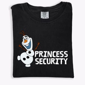 May include: Black t-shirt featuring a cartoon image of Olaf the snowman with the text "PRINCESS SECURITY" in white. The shirt is folded and has a Comfort Colors tag at the top. The design is centered on the front of the shirt.