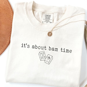 Pu&ograve; includere: T-shirt color crema con la scritta nera "it's about bam time". La maglietta presenta una grafica di due tessere di mahjong. Una collana d'oro con un ciondolo &egrave; drappeggiata sulla maglietta.